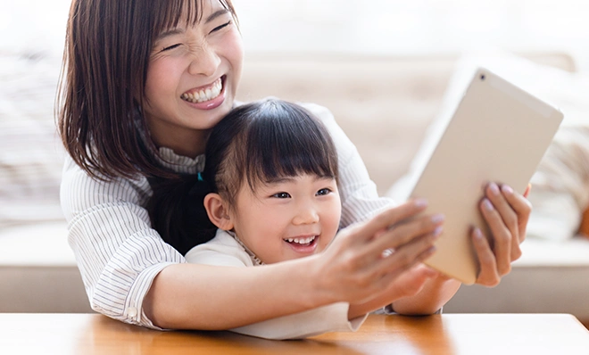 タブレットを手に持ち、満面の笑みで画面を覗き込む母親と、その腕の中で楽しそうにしている小さな女の子